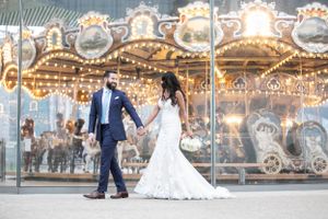 A couple shares a romantic moment in front of an illuminated vintage carousel at dusk, the bride wearing a flowing lace gown.