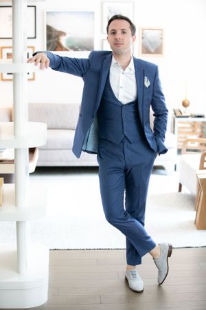 A person in a stylish navy blue three-piece suit and white dress shoes leans casually against a white pillar in a modern interior.