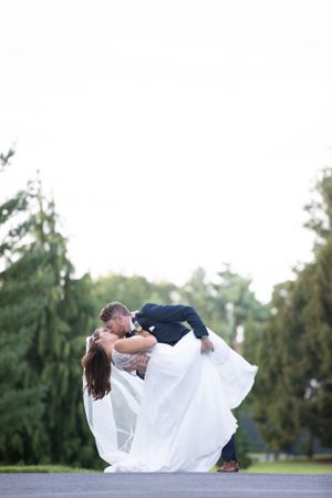 A romantic couple in elegant wedding attire share a dramatic dip pose on their special day surrounded by evergreen trees.