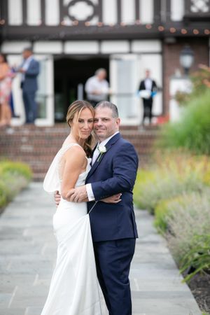 A bride in a sleek white dress and groom in navy suit embrace on a garden pathway near a brick building.