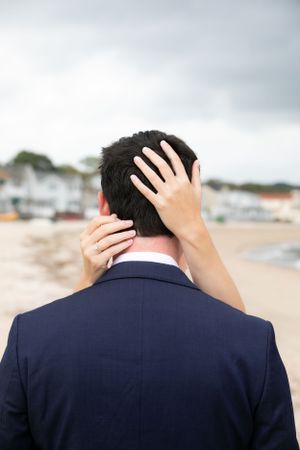 Back view of person in navy suit with hands placed gently on their neck and head, standing on a beach.