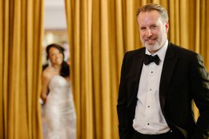 A groom in a black tuxedo stands smiling with gold curtains in the background while a bride is visible in soft focus.