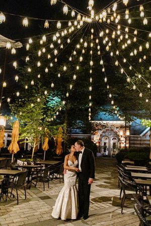 A romantic evening scene with string lights illuminating an outdoor patio with tables and chairs near an ornate building entrance.
