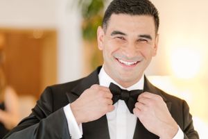 A well-dressed person in a black tuxedo and bow tie adjusts their collar while smiling warmly.