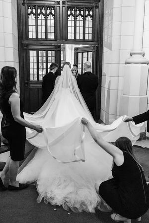 Wedding assistants help adjust an elegant white wedding gown and long flowing veil at Gothic church doorway entrance.