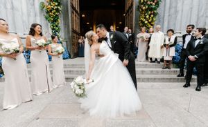 A couple shares a romantic wedding kiss on church steps surrounded by bridesmaids in blush dresses and groomsmen.