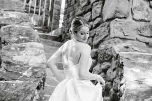 A black and white photograph shows a bride in a strapless wedding dress seated on stone steps beside a rock wall.