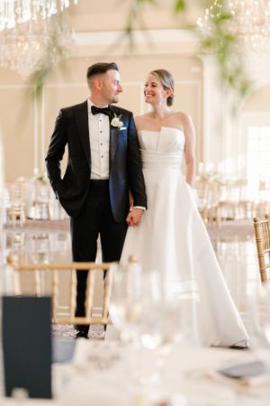 A happy wedding couple stands together in an elegant ballroom with gold chairs and soft decor.