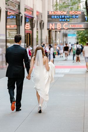 A couple in wedding attire walks down a city sidewalk near NBC Studios with the Rainbow Room sign visible above.