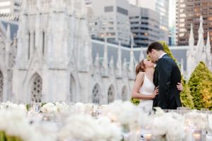 A romantic wedding couple embraces outside a majestic cathedral with white flowers in the foreground.
