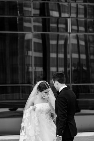A bride in a delicate lace veil and groom in black suit share an intimate moment outside a modern glass building.