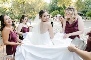 A group of bridesmaids in burgundy dresses help adjust a bride's flowing white wedding dress outdoors.