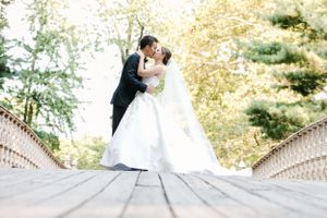 A couple in wedding attire share a romantic moment on a wooden bridge surrounded by sunlit trees.