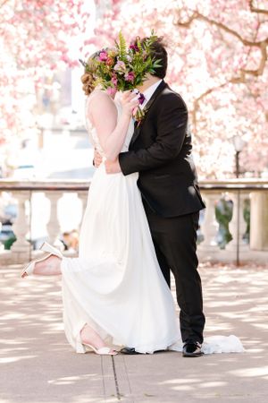 A romantic wedding photo under pink cherry blossoms with the bride in white and groom in classic black suit.