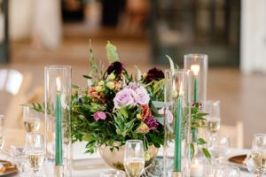Elegant wedding table centerpiece with purple roses, greenery and tall glass candle holders on a formal dining setting.