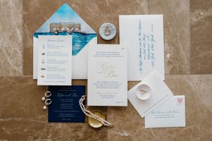Wedding invitation suite in navy blue and white with gold rings and feather accent displayed on marble surface.