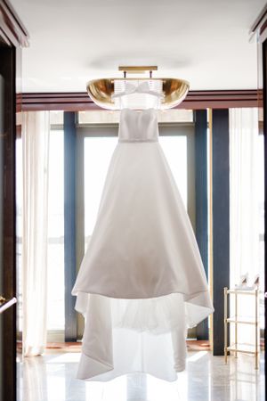 A white wedding dress hangs elegantly against a window, with natural light streaming in to illuminate its classic silhouette.