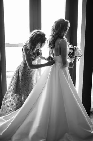 A black and white wedding photograph shows a tender moment as someone helps adjust an elegant ballgown wedding dress.