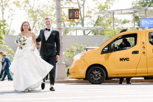 A couple in wedding attire walks past a yellow NYC taxi on a sunny city street.