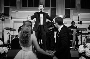 Black and white photo of a wedding reception with a performer singing on stage while a bride and groom watch from below.