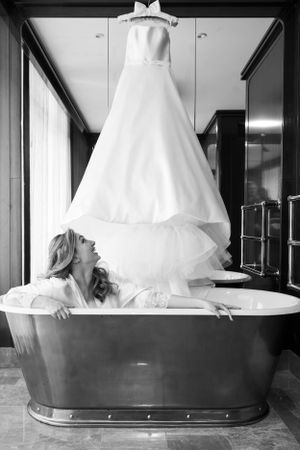 Black and white photo showing a white wedding dress hanging above a freestanding bathtub in an elegant bathroom setting.