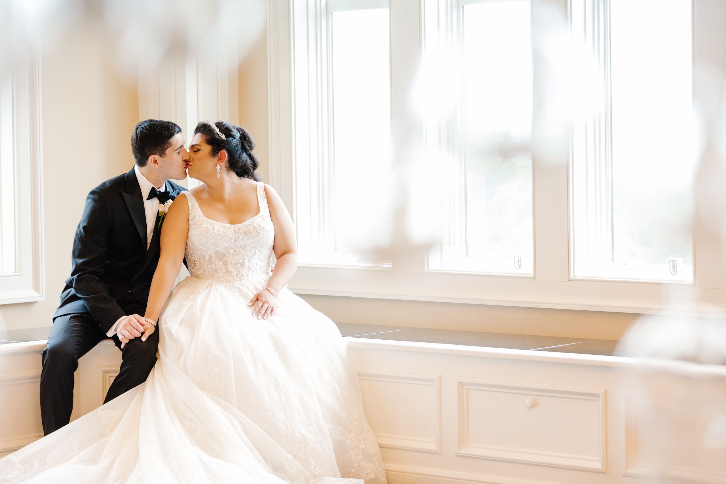 A couple shares an intimate moment together in a bright sunlit room while wearing formal wedding attire.