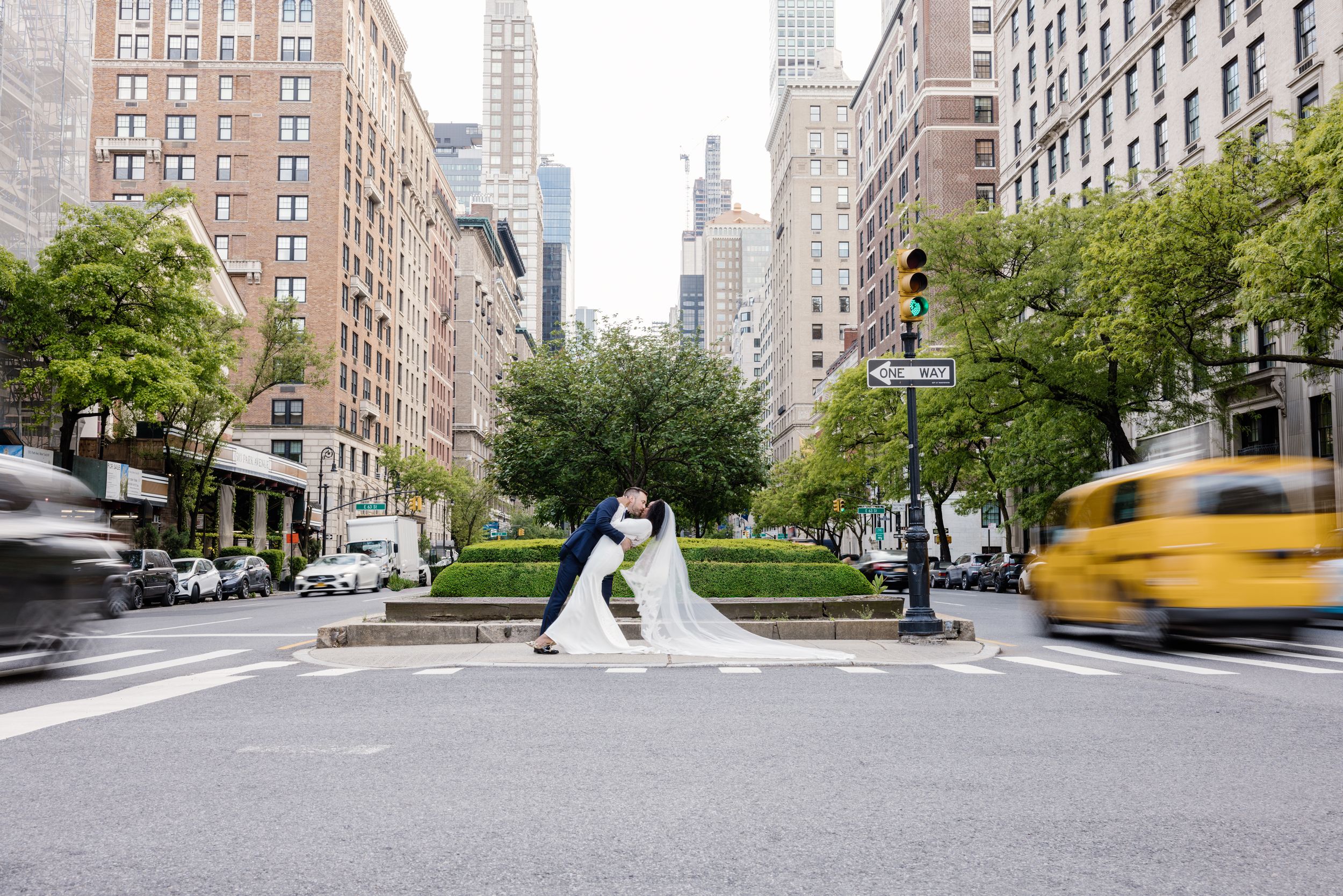Two figures pose on a city street in New York with tall buildings and a yellow taxi blurring past in motion.