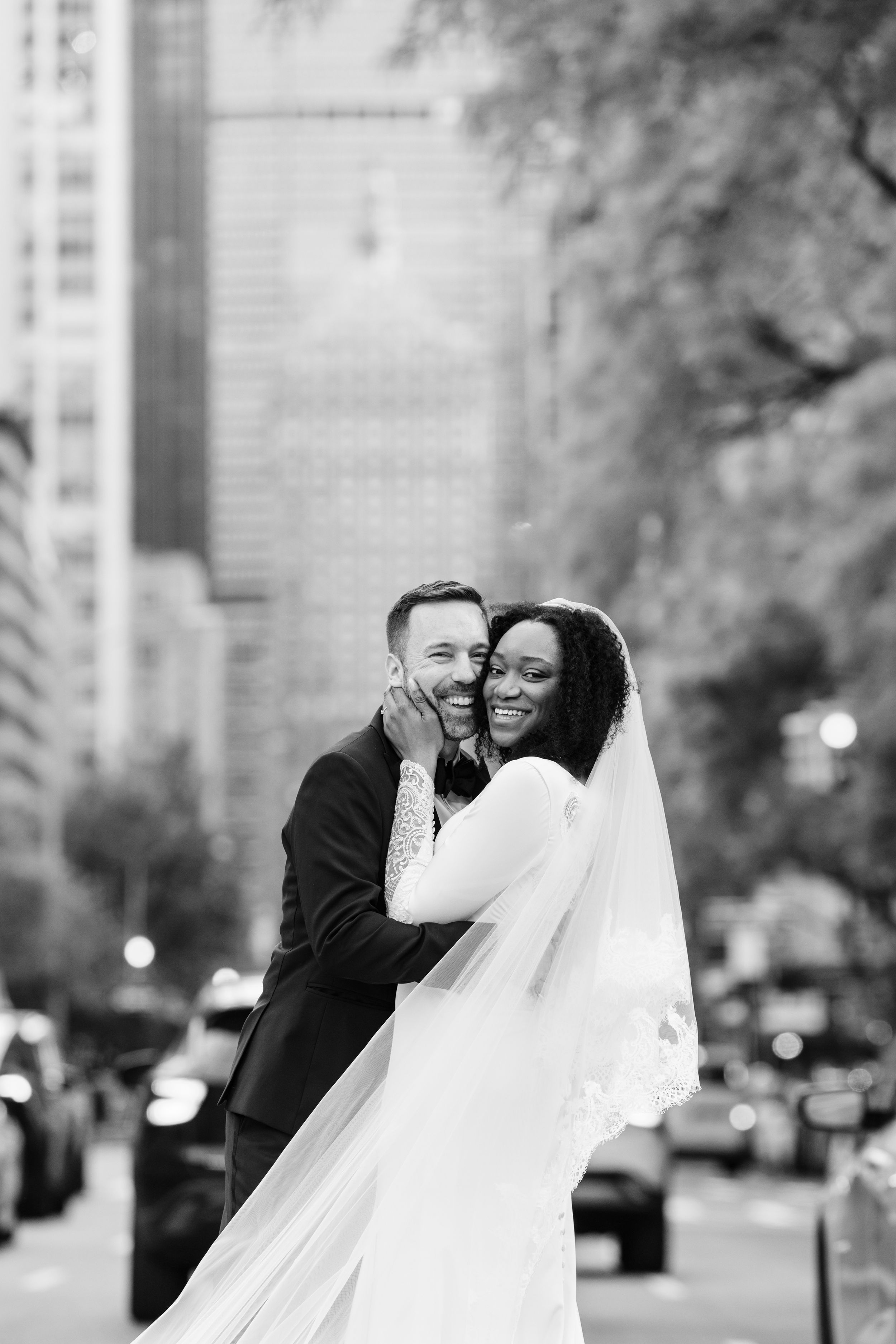 A happy couple shares a tender moment on their wedding day in an urban setting with city buildings in the background.