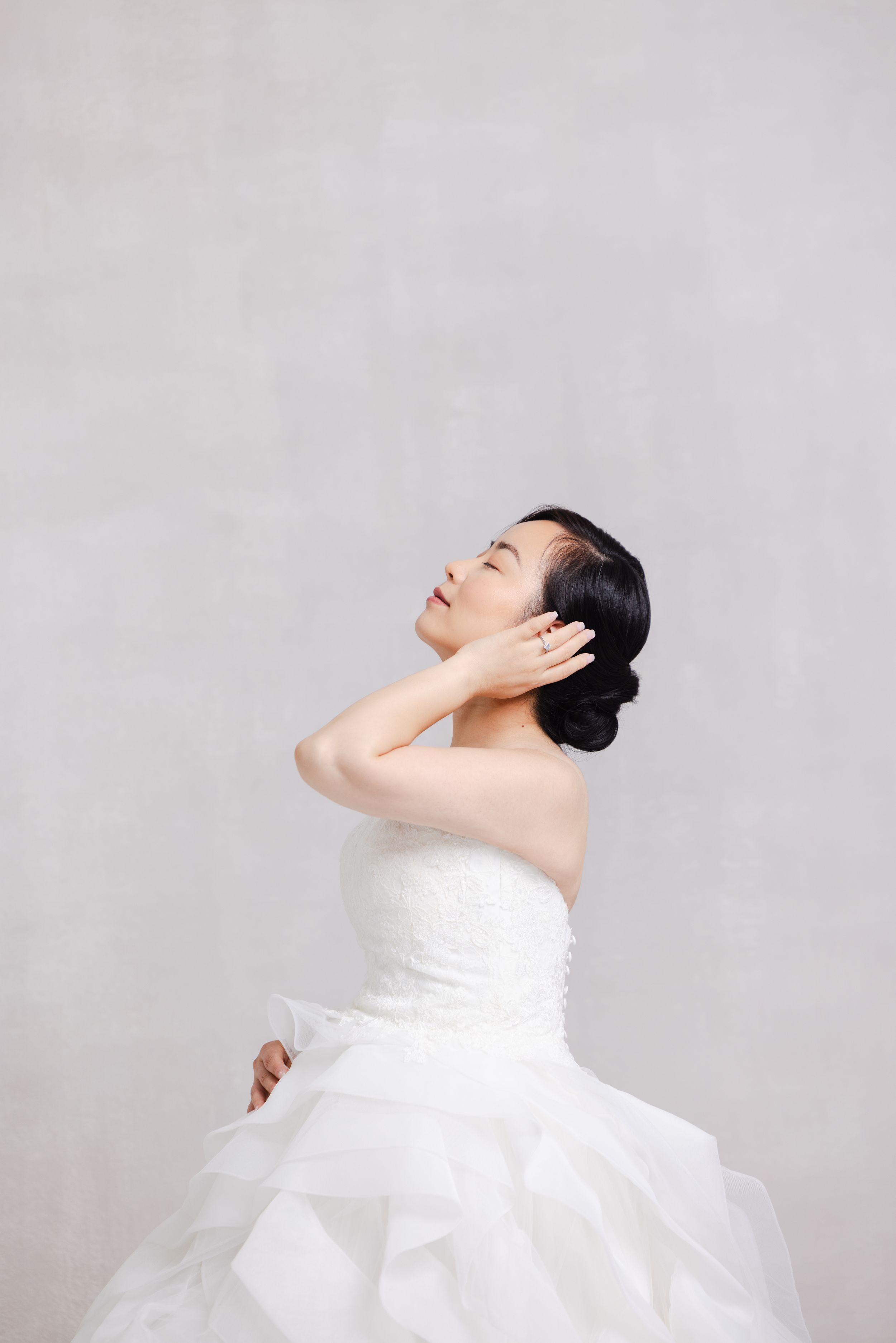 A bride in a white strapless ballgown holds her hands gracefully near her neck while looking up against a gray background.