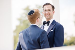Two people in navy blue tuxedos share a joyful moment at a formal wedding ceremony outdoors.