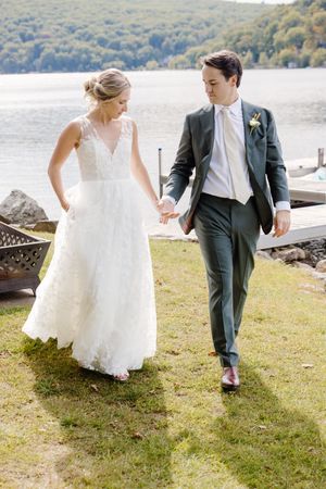 A couple in wedding attire walk together on a grassy lakeside shore, with mountains and water in the background.