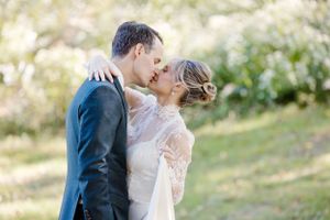 A romantic wedding couple embraces and kisses in an outdoor garden setting with soft natural sunlight.