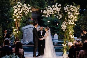Beautiful outdoor wedding ceremony under an illuminated floral arch with white orchids at dusk.
