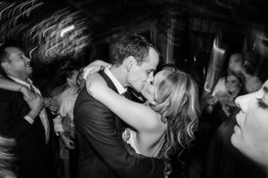 A romantic moment of a couple kissing on a crowded dance floor at what appears to be a wedding reception in black and white.