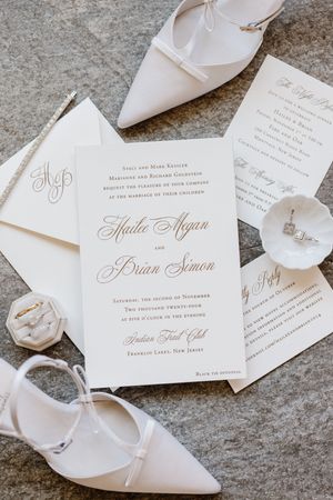 Elegant white wedding invitation suite with calligraphy text layout alongside matching white pointed-toe bridal shoes.