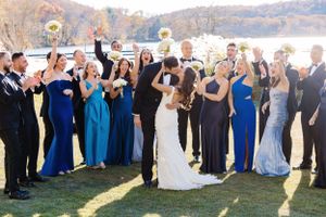 Wedding party celebrates outdoors with raised hands in blue and black attire by scenic waterfront on sunny day.