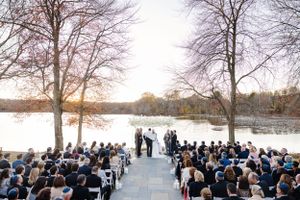 Wedding ceremony taking place at sunset by a lake with guests seated in rows and bare trees framing the waterfront scene.