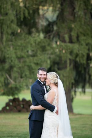 A bride and groom embrace outdoors under evergreen trees during their wedding celebration.