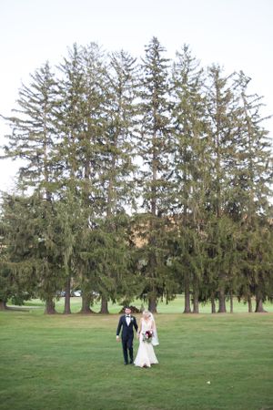 A couple walks together across a lush green lawn with tall evergreen trees creating a stunning natural backdrop.