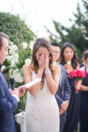 Bride in white lace dress covers her face in an emotional moment during outdoor wedding ceremony among flowers.