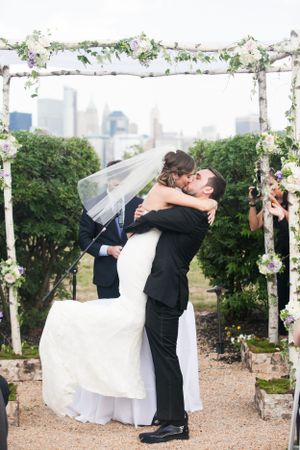 A romantic kiss under a floral arch decorated with white flowers and greenery, with a city skyline visible in the background.