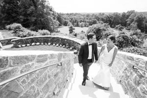 A couple in formal wedding attire walks up stone steps at an outdoor venue with a scenic overlook.