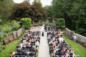 Outdoor wedding ceremony taking place in a garden courtyard surrounded by stone walls and lush greenery.