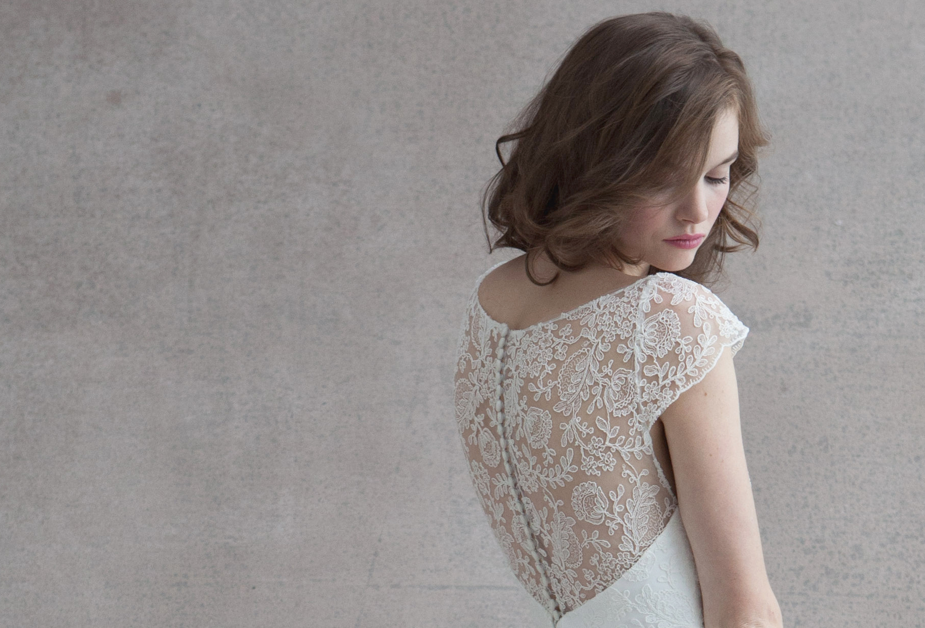 Elegant white lace wedding dress with delicate sheer back detail shown against neutral grey background.