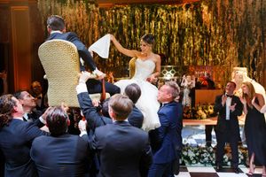 Bride and groom lifted in chairs during lively Jewish wedding hora dance celebration with guests cheering.