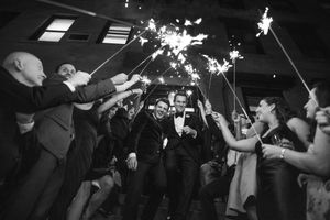Black and white photo of two grooms walking through a sparkler tunnel surrounded by cheering wedding guests at night.