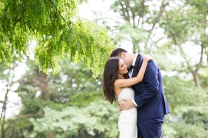 A romantic couple shares a tender moment while embracing under lush green trees in a garden setting.