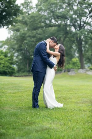 A romantic couple in wedding attire share an intimate embrace on a lush green lawn with trees in the background.