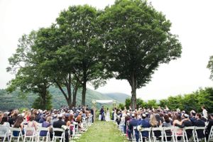 Outdoor wedding ceremony setup with white chairs on a hilltop under large green trees on a sunny summer day.