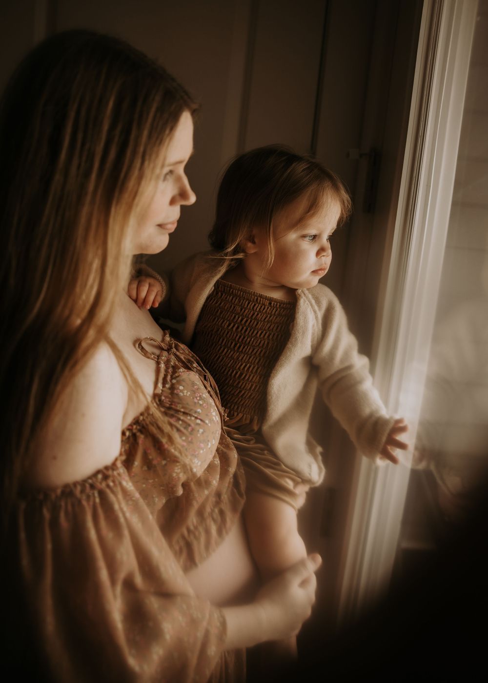 Two figures stand by a sunlit window in a warm, sepia-toned scene wearing light colored clothing.
